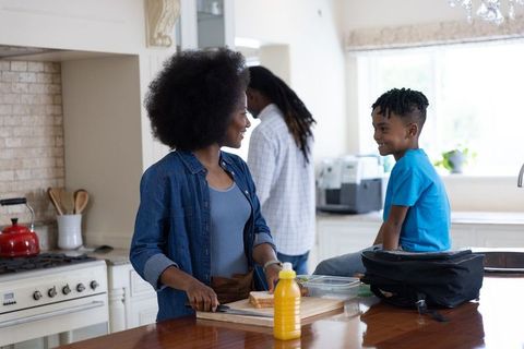 African american mom preparing school lunch while son sits on counter smiling
