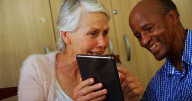 A senior Caucasian woman and an African American man share a joyful moment looking at a tablet, with copy space. Their warm interaction suggests they are enjoying a digital experience together, connecting with family or friends online.