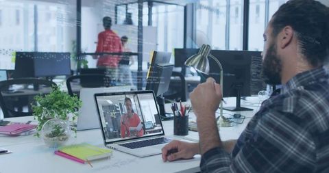 Bearded man gesturing during video call at modern open office workstation with laptop teleconference