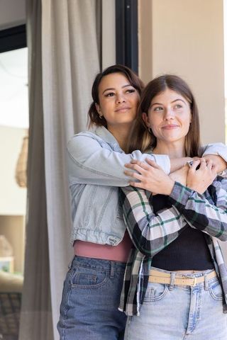 Female partners embracing in home doorway, smiling and hugging, casual denim and plaid