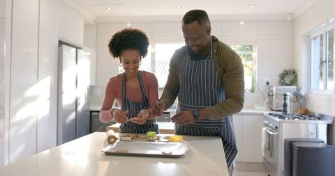 Smiling couple baking cookies on bright modern kitchen island wearing matching aprons