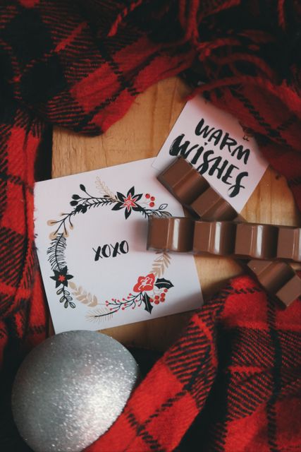 Festive holiday scene includes greeting cards with 'Warm Wishes' and 'XOXO' messages, red and black plaid fabric, chocolates and a silver orb decoration. Perfect image for season's greetings, festive promotions, and holiday-themed social media posts.