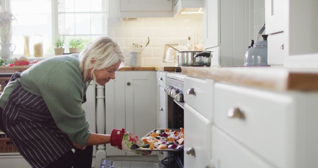 Senior woman placing fresh vegetables into oven for baking in bright home kitchen. Great for themes related to healthy lifestyle, wellness, home cooking, elderly activities, and wholesome eating. Suitable for use in articles, blogs, advertisements promoting healthy habits and senior living.