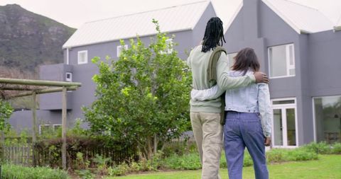 Diverse couple walking toward modern two-story house with lush garden and pergola