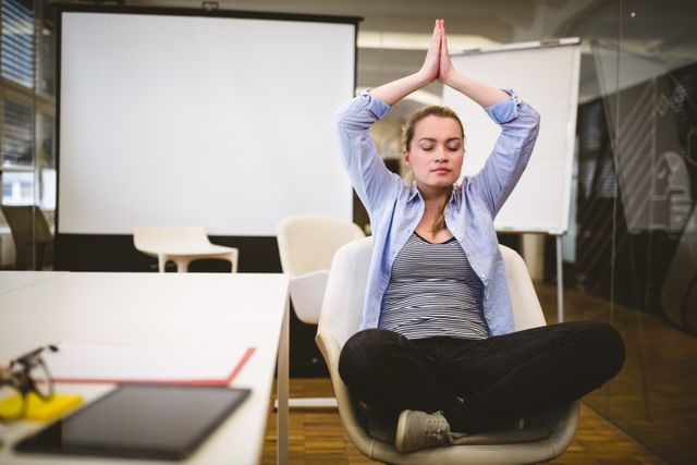 Young businesswoman practicing yoga in an office meeting room, promoting workplace wellness and stress relief. Ideal for use in articles or presentations about corporate wellness programs, mindfulness at work, and maintaining work-life balance.