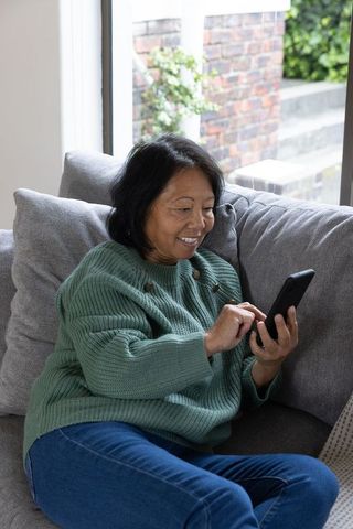Senior Asian Woman Smiling Using Smartphone on Sofa by Window