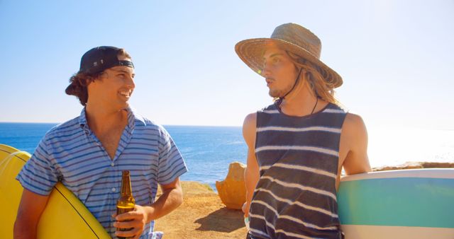 Two young men standing on a beach with ocean view, each holding a surfboard and relaxing. One man wears a striped tank top and sun hat, while the other wears a striped shirt and baseball cap, holding a beer bottle. Perfect for content about surfing lifestyle, beach activities, summer tourism, and male friendships.