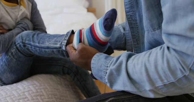 Parent assisting child with putting on striped socks while both wearing denim jeans. Captures a moment of care, intimacy, and everyday life at home. Suitable for themes related to parenting, family support, and nurturing. Could be used in family-centered advertisements, articles on parenting tips, or content illustrating daily family interactions.