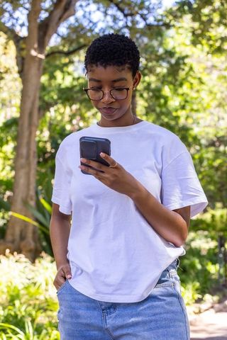 African woman standing in sunny park wearing white tee and jeans texting on smartphone