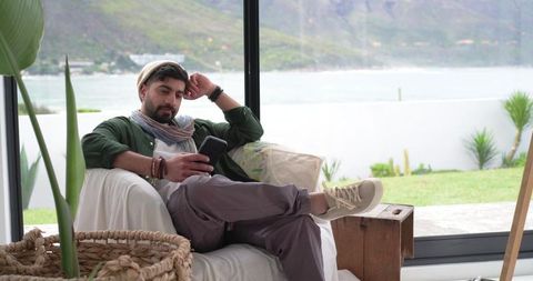 Indian man relaxing on coastal living room sofa using smartphone, modern casual lifestyle