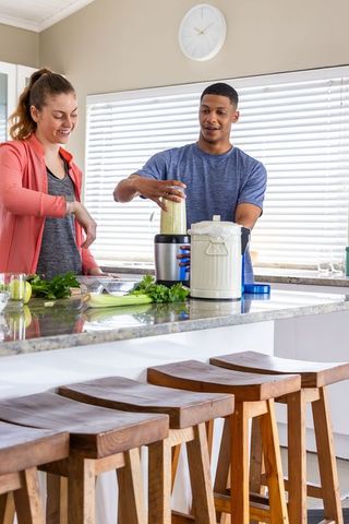 Couple preparing green smoothie at bright kitchen island, blending healthy drink together