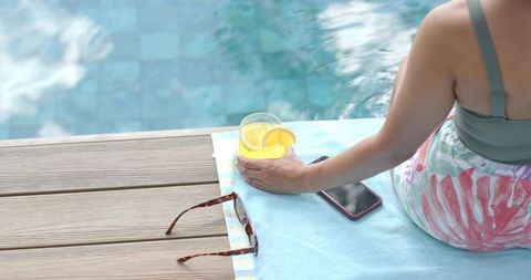 African american woman sitting poolside holding orange drink and phone, summer relaxation