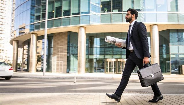 Confident businessman walking with briefcase and rolled newspaper past glass office tower