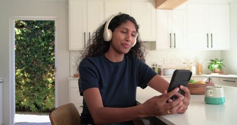 African american woman wearing headphones and texting on smartphone at kitchen island