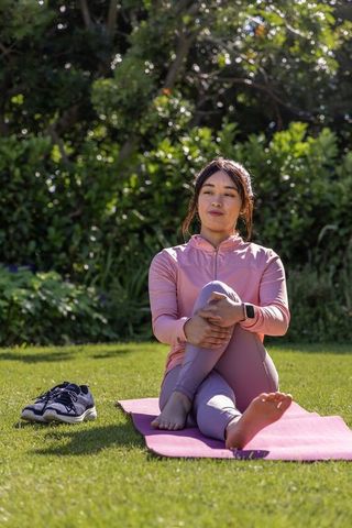 Asian woman stretching on pink yoga mat in garden wearing smartwatch barefoot wellness