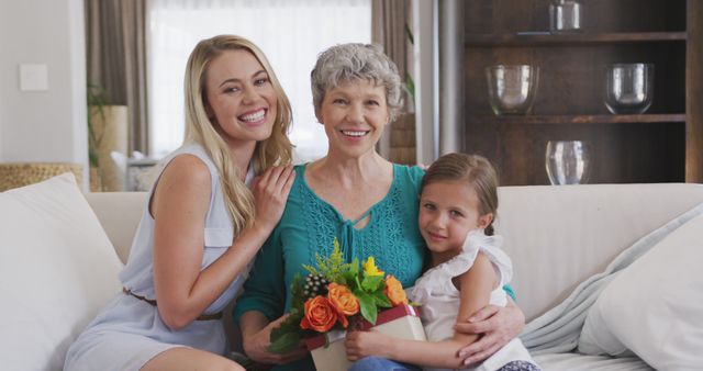 Portrait of happy caucasian grandmother, mother and daughter sitting on sofa, and holding present. Mother's day, celebration, domestic life, family, and togetherness.