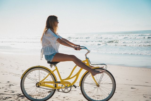 Caucasian woman enjoying a sunny day by the sea, riding a yellow bicycle on the sandy beach. Ideal for promoting summer vacations, outdoor activities, travel destinations, and healthy lifestyles. Perfect for use in advertisements, travel brochures, and lifestyle blogs.
