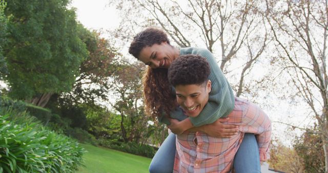 This image shows a happy couple laughing and enjoying a playful piggyback ride in a park on a sunny day. Ideal for use in advertisements, blog posts, and articles about relationships, outdoor activities, or lifestyle content focusing on happiness and togetherness.