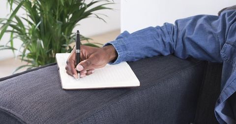African American man writing in lined notebook on sofa armrest wearing denim shirt