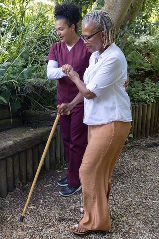 Caregiver guiding senior african american woman with cane on sunlit garden path