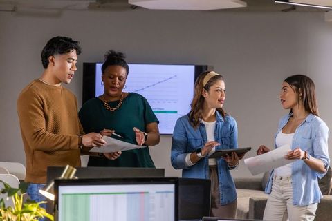 Diverse Team Collaborating Over Reports and Tablet in Open Office With Analytics Display
