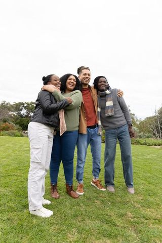 Diverse friends laughing and hugging on park lawn wearing casual jackets and scarves
