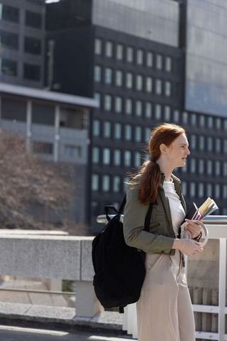 Redheaded woman standing on city bridge holding notebooks and pen carrying black backpack