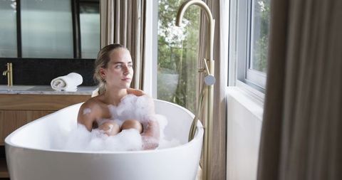 Young woman relaxing in freestanding bathtub with bubbles, gazing out window at forest view
