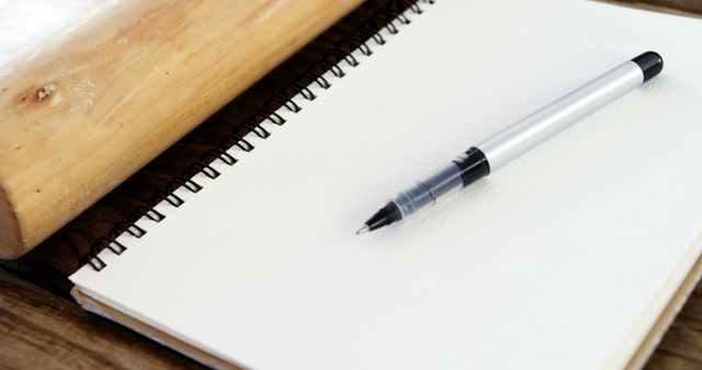 A pen lies on a blank spiral notebook next to a wooden rolling pin, with copy space. It suggests a fusion of cooking and recipe noting or the planning of a baking project.
