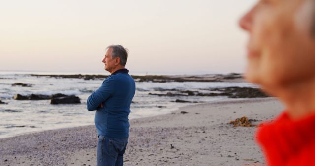 Older couple is standing on a calm beach at sunset, with an ocean backdrop and sandy shore. Ideal for use in articles or advertisements focusing on retirement, senior lifestyles, relaxation, travel, romantic getaways, or serenity.