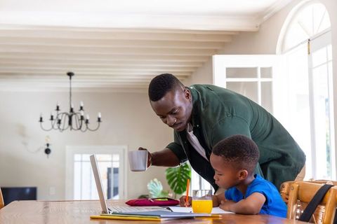 African american father helping son with homework at bright dining table with laptop