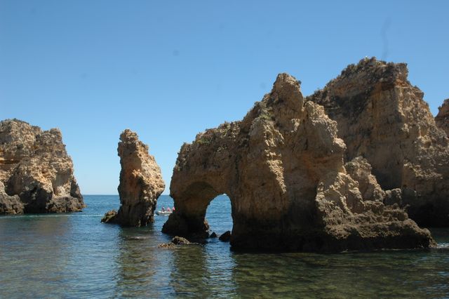 Rock formations and cliffs located along an ocean coastline under a clear blue sky. The natural arch and tranquil waters create a picturesque view. Ideal for use in travel brochures, nature publications, wallpapers, and landscape photography collections.