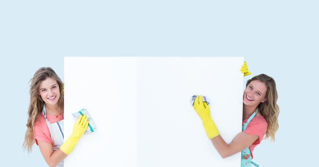 Two women smiling while cleaning with gloves and cloths. Ideal for advertisements or promotions related to household chores, cleaning services, teamwork, and positive lifestyle themes.