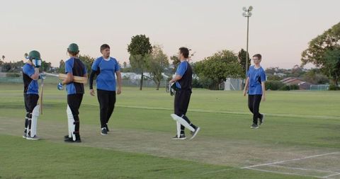 Regrouping male cricket players on pitch at dusk wearing blue jerseys and protective gear
