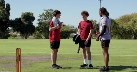 Three male cricket players chatting on sunny green pitch holding helmet and batting gear