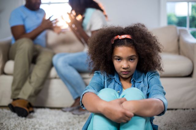Young girl sitting on floor with a sad expression while parents argue in the background. Useful for illustrating family conflict, emotional distress in children, and the impact of domestic issues on young ones. Ideal for articles, blogs, and educational materials on family dynamics, child psychology, and mental health.