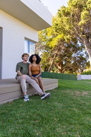 Young couple relaxing on raised deck of modern white home with lush backyard lawn