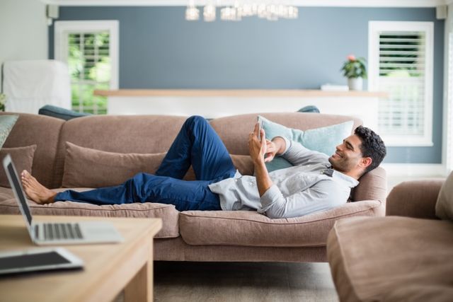 Man lying on a comfortable sofa using a mobile phone in a modern living room. Ideal for depicting relaxation, leisure time, home lifestyle, and modern technology use. Suitable for articles or advertisements related to home decor, technology, or lifestyle.