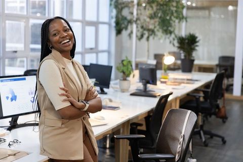 Confident African American businesswoman smiling while leaning on desk in open office