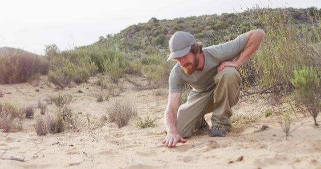 Caucasian male survivalist in wilderness squatting down and measuring animal print in sand with hand. exploration, travel and adventure, survivalist in nature.