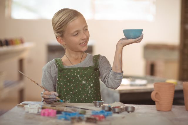 Young girl in pottery workshop holding and admiring a handmade bowl. She is smiling and appears proud of her creation. The scene is filled with various pottery tools and paints, indicating an active and creative environment. This image is perfect for use in educational materials, art and craft promotions, children's activities, and creative hobby advertisements.