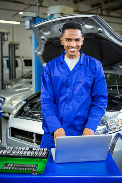 Young mechanic in blue jumpsuit using a laptop in an auto repair shop, smiling confidently. Ideal for use in automotive industry advertisements, mechanic training materials, technology in car repair contexts, and professional service promotions.