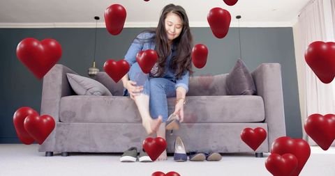Asian woman putting on shoes on sofa surrounded by floating red heart balloons