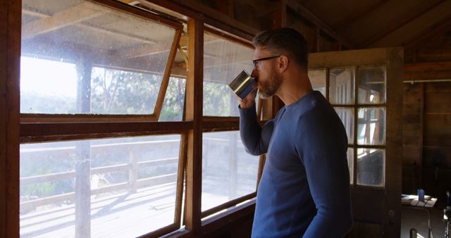 Man enjoys his morning coffee while looking out of a rustic cabin window. Ideal for themes about relaxation, nature getaways, leisure time, and peaceful environments.