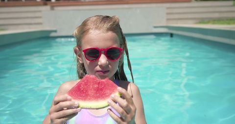 Preteen girl eating watermelon in pool wearing red sunglasses and one-piece swimsuit