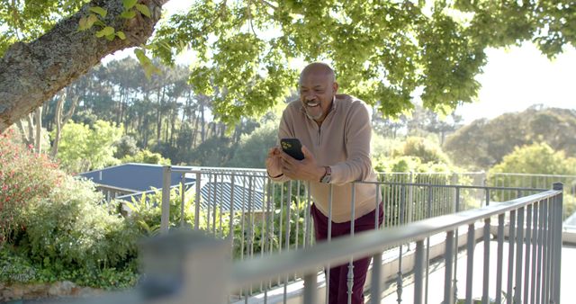 This image depicts a man talking on a smartphone while leaning on a railing outdoors, surrounded by lush green trees. He is smiling, indicating a pleasant conversation. This picture is ideal for illustrating concepts of modern communication, people enjoying technology, outdoor activities, and positive emotions. Potential uses include promotional materials for mobile carriers, tech gadgets, or lifestyle blogs focused on technology and nature.