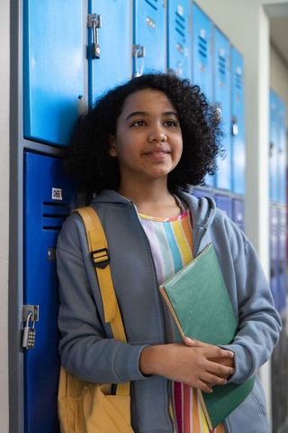 African american teen leaning on blue lockers holding green binder and backpack