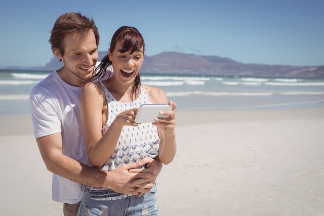 Couple enjoying a sunny day at the beach while looking at a mobile phone. Perfect for themes related to summer vacations, technology use in outdoor settings, relationships, and leisure activities. Ideal for travel blogs, social media posts, and advertisements promoting beach destinations or mobile technology.