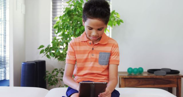 Boy sitting on couch holding tablet, focused on screen. Wearing orange striped shirt, modern living room with natural light and plants. Ideal for technology use by children, digital learning, home environments, casual lifestyle, and modern family settings.