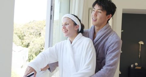 Sunlit morning romance with diverse couple embracing at open window in cozy bathrobes